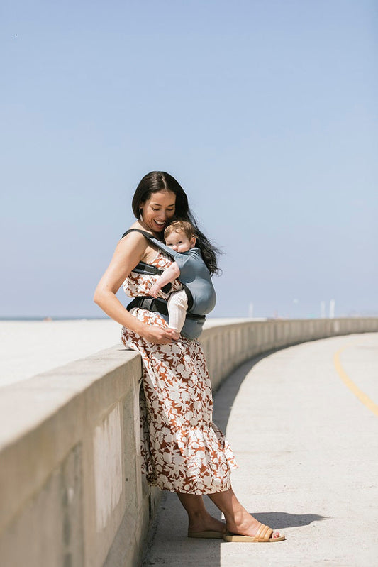 Mama nosi dziecko przodem do rodzica na plaży w lekkim nosidełku podróżnym Tula Lite.