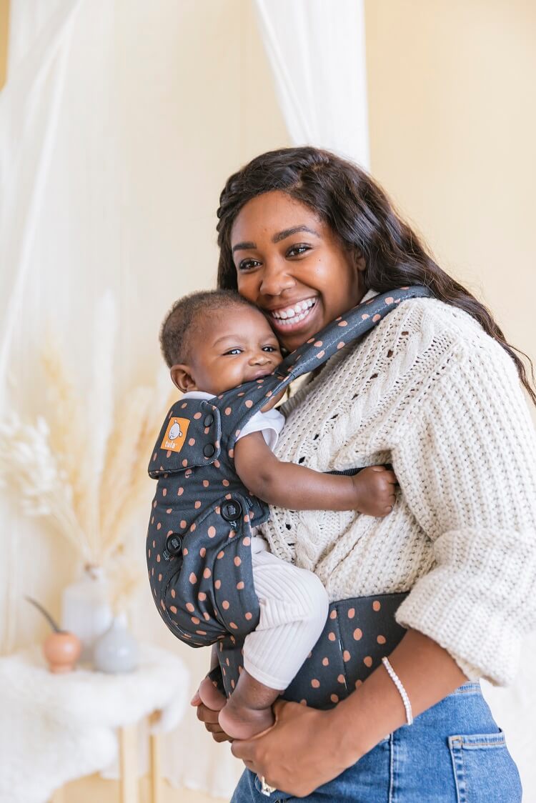 Uśmiechnięta mama nosi wesołe dziecko w nosidełku Tula Explore Ginger Dots z bawełny