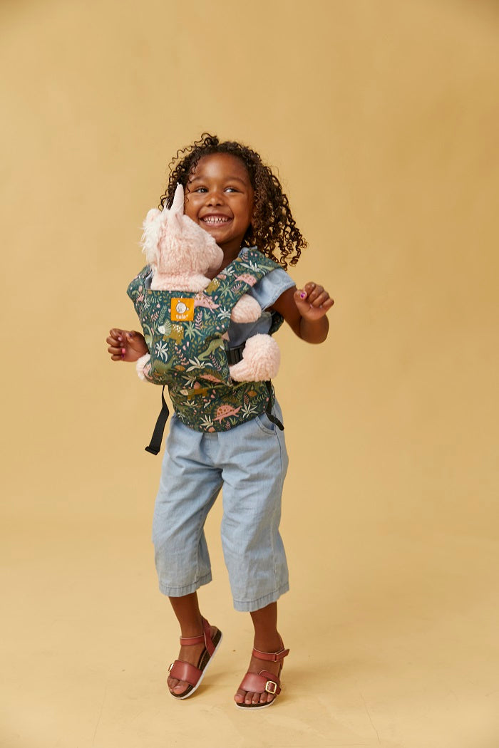A girl jumping and carrying her unicorn toy in Tula Mini Doll Carrier in fun dinosaur print
