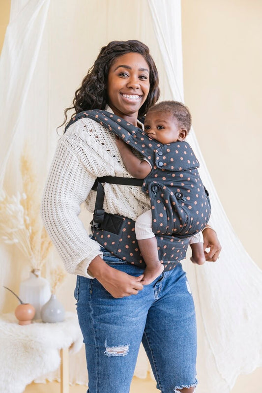 Uśmiechnięta mama nosi niemowlaka w nosidełku Tula Explore Ginger Dots z bawełny w ergonomicznej pozycji