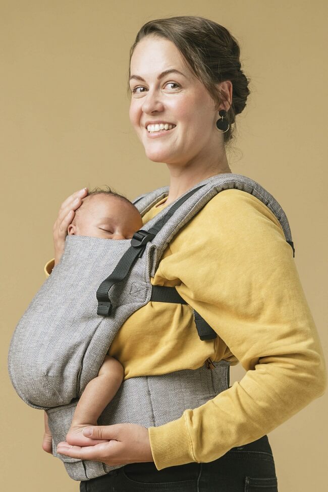 Uśmiechnięta mama tuli niemowlaka w pozycji przodem do rodzica w nosidełku Tula Linen Free-to-Grow Ash
