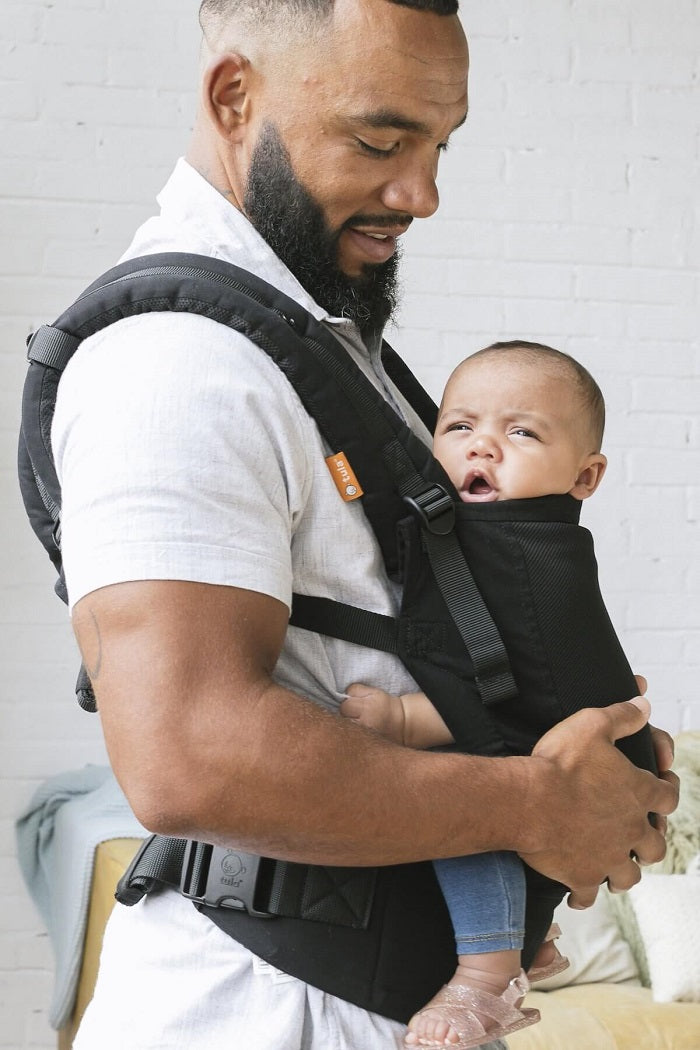 Tata nosi malutkie dziecko w nosidełku Tula Free-to-Grow Coast Urbanista w neutralnym czarnym kolorze z wstawką z przewiewnej siateczki