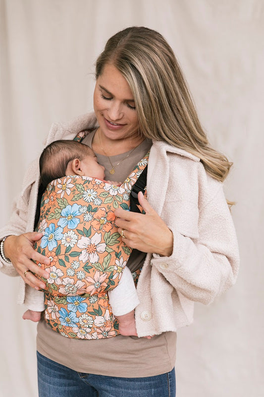 mama nosi dziecko w ergonomicznej pozycji w nosidełku Tula Free-to-Grow Flower Walk z wzorem kwiatów na pomarańczowym tle