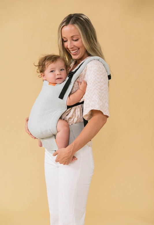 Uśmiechnięta mama nosi zaciekawione dziecko w ergonomicznej pozycji w nosidełku Tula Linen Free-to_grow Seafoam