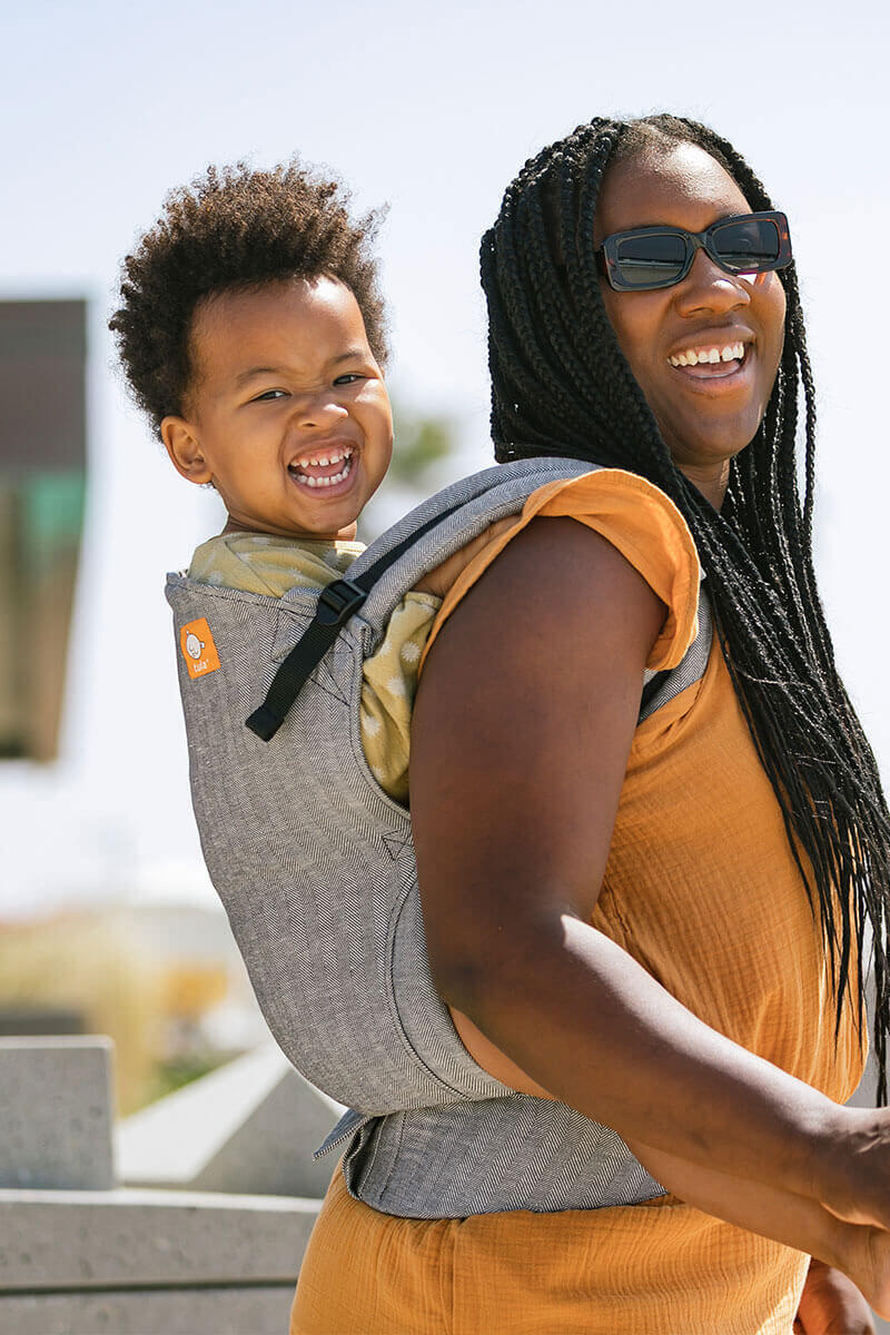 Roześmiana mama nosi zadowolonego przedszkolaka w nosidełku Tula Toddler