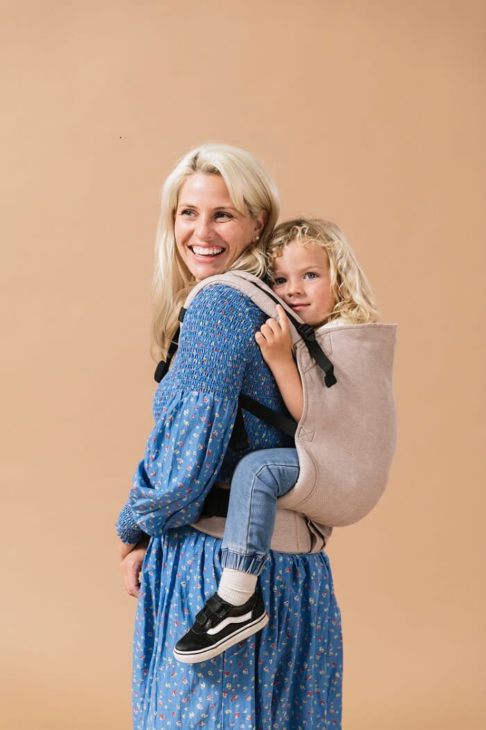 Mama nosi dziecko na plecach w nosidełku Tula Toddler Linen Sand.