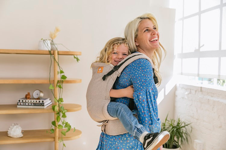 Uśmiechnięta mama nosi dziecko na plecach w nosidełku Tula Toddler Linen Sand.