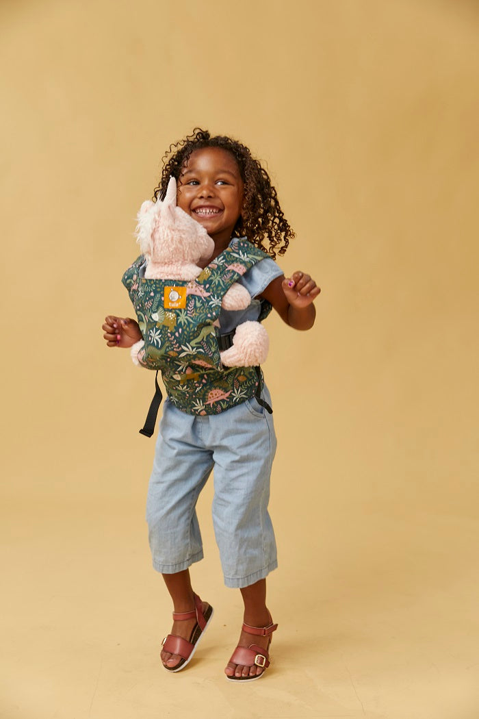 A girl jumping and  carrying her unicorn toy in Tula Mini Doll Carrier in fun dinosaur print